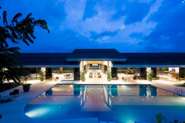 A luxurious pool and modern open-air building are illuminated at night, reflecting on the water under a blue, slightly cloudy sky.