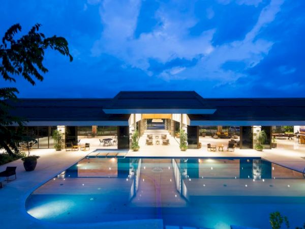 A luxurious pool and modern open-air building are illuminated at night, reflecting on the water under a blue, slightly cloudy sky.