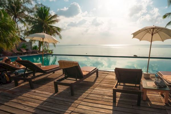 A tropical poolside with lounge chairs, umbrellas, palm trees, and a view of the ocean under a partly cloudy sky.