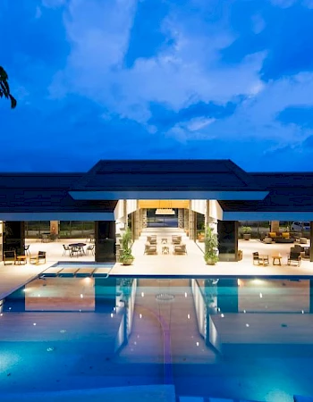 The image shows a luxurious outdoor swimming pool area by a modern building, illuminated at dusk with a serene atmosphere.