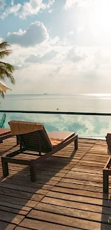 A serene poolside setting with lounge chairs, umbrellas, and a view of the ocean, surrounded by palm trees under a bright sky.