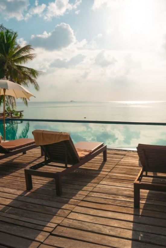 Poolside scene with lounge chairs, umbrellas, palm trees, and ocean view under a cloudy sky.