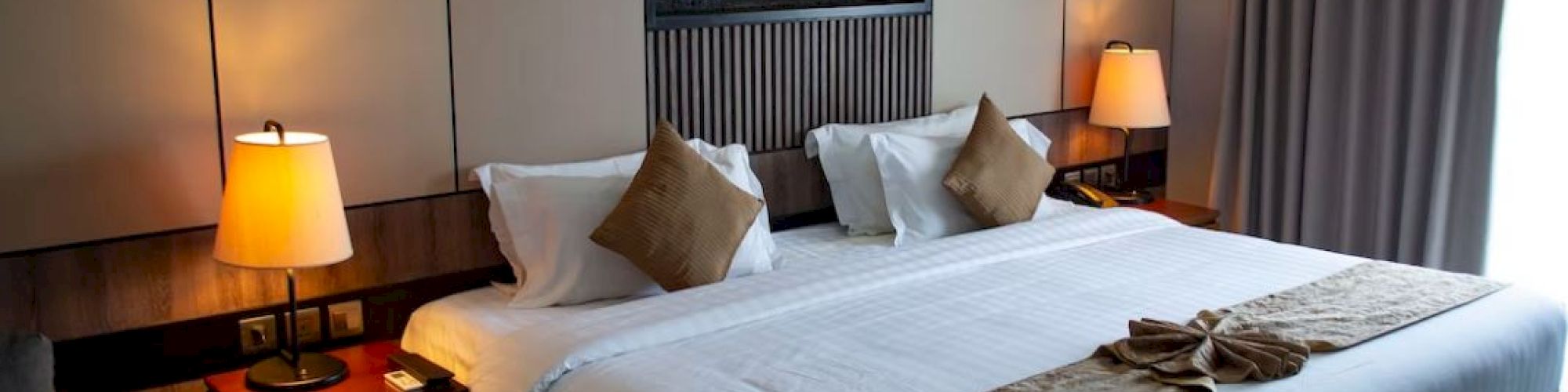 The image shows a neatly arranged hotel room with a large bed, two bedside lamps, decorative pillows, and a wall art piece above the bed.
