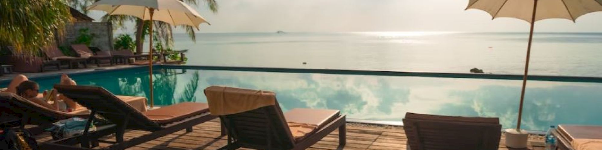 A serene poolside scene with lounge chairs, umbrellas, and a view of calm water under a sunny sky, surrounded by palm trees.