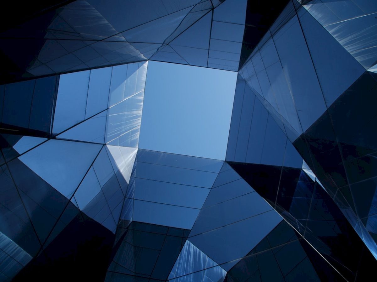 The image shows a geometric pattern of reflective glass buildings framing a patch of blue sky, creating a symmetrical architectural view.
