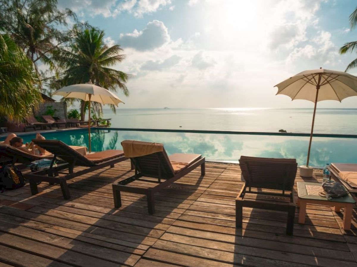 A serene poolside with deck chairs, umbrellas, palm trees, and a view of the ocean under a partly cloudy sky at sunrise or sunset.