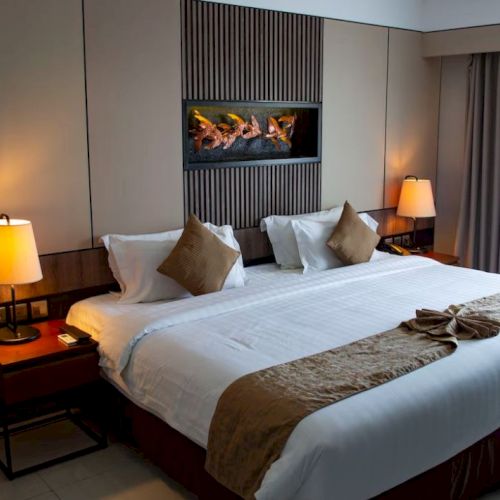 A neatly arranged hotel room with a king-size bed, plush pillows, bedside lamps, a small table, and a decorative wall feature above the bed.