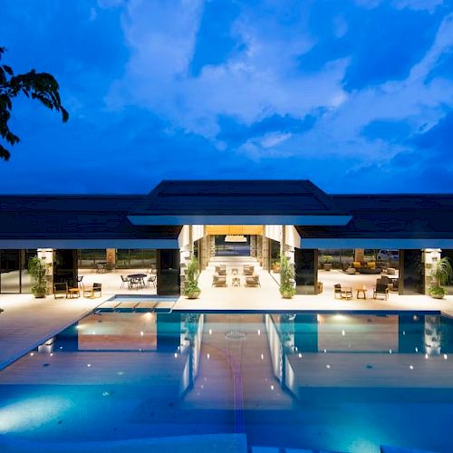 A luxurious modern building with a large illuminated swimming pool at dusk, surrounded by seating areas and trees.