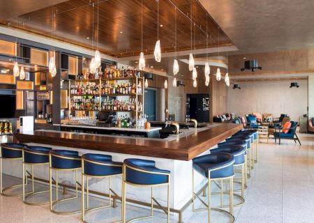 This image shows a modern bar with blue stools, a wooden counter, shelves of drinks, and stylish lighting in a spacious lounge setting.