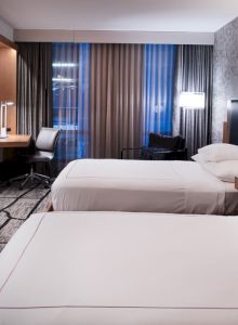 A modern hotel room with two beds, TV, desk, and large windows, featuring a stylish décor and lighting.