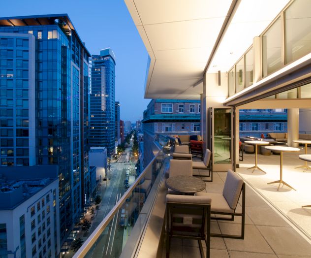 A modern rooftop terrace at dusk, overlooking a city street with tall buildings. The terrace has tables and chairs along the railing.