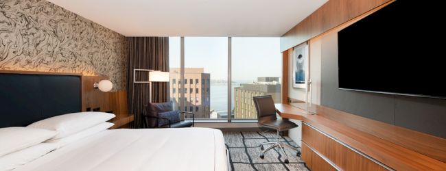 A modern hotel room with a large bed, TV, desk, and chair. The window offers a view of nearby buildings and water in the distance.