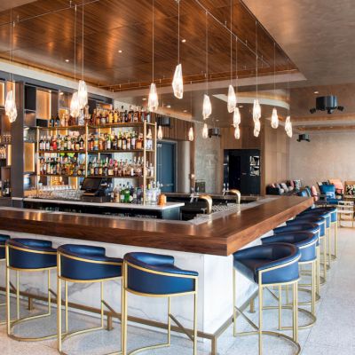A modern bar area with a long counter, bar stools, and shelves stocked with various liquors. Elegant lighting hangs from the ceiling.