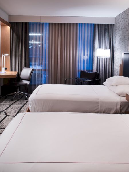 The image shows a modern hotel room with two beds, a flat-screen TV, a desk, and a large window with curtains.