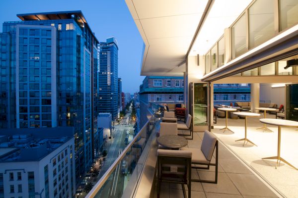 A modern outdoor balcony overlooking a cityscape at dusk, with seating and tables, adjacent to a glass-walled indoor area.