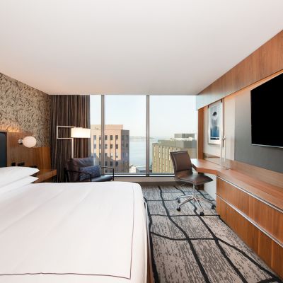 A modern hotel room with a large bed, desk, chair, and TV. Large window offers a city view. Decorative wall with neutral tones.