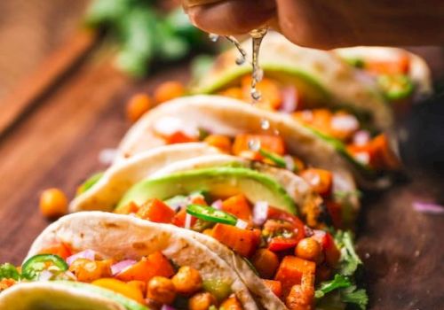 The image shows tacos filled with chickpeas, avocado, and vegetables on a wooden board, with lime being squeezed over them.