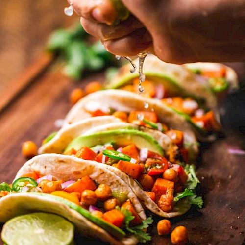The image shows tacos filled with chickpeas, avocado, and vegetables on a wooden board, with lime being squeezed over them.
