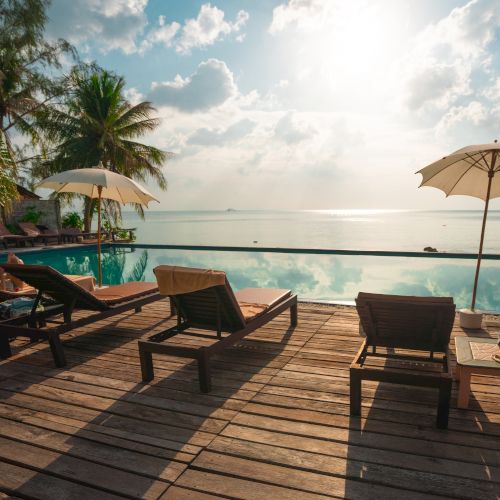A scenic view of a poolside area with lounge chairs, umbrellas, and palm trees overlooking a peaceful ocean under a sunny sky.