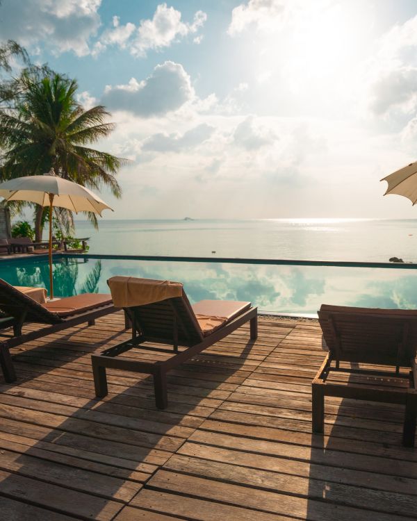 A scenic view of a poolside area with lounge chairs, umbrellas, and palm trees overlooking a peaceful ocean under a sunny sky.