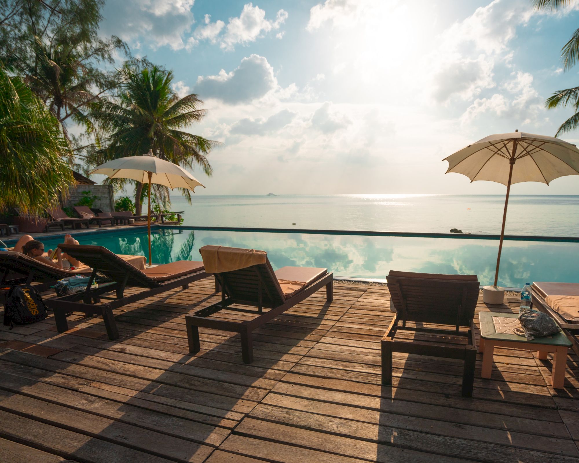 Deck with lounge chairs and umbrellas by a pool overlooking a calm ocean, surrounded by palm trees under a partly cloudy sky.