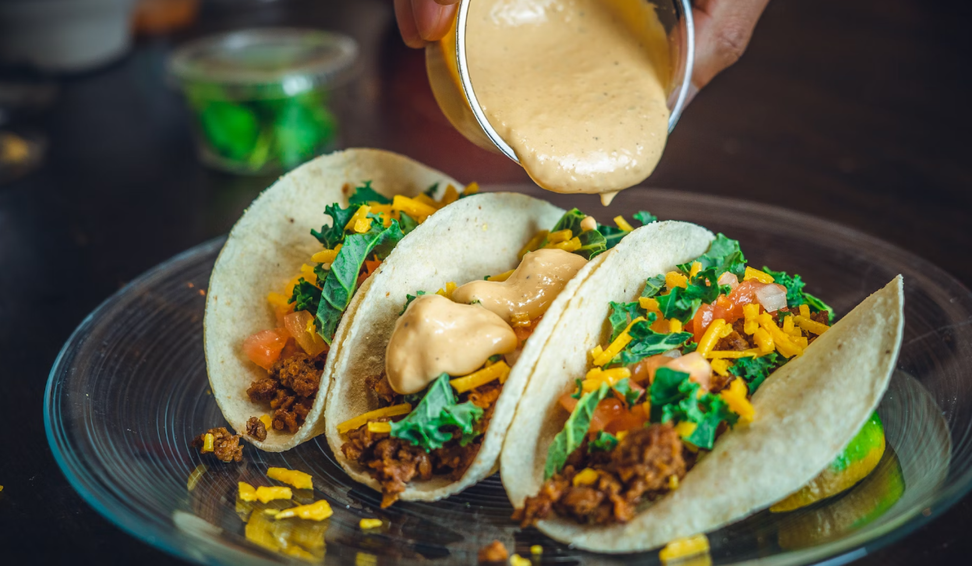 Three tacos filled with ground beef, cheese, lettuce, and tomatoes are being topped with creamy sauce on a glass plate.