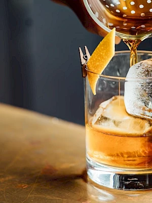 A cocktail is being poured into a glass with a large ice cube and a twist of lemon peel garnish, sitting on a smooth surface.