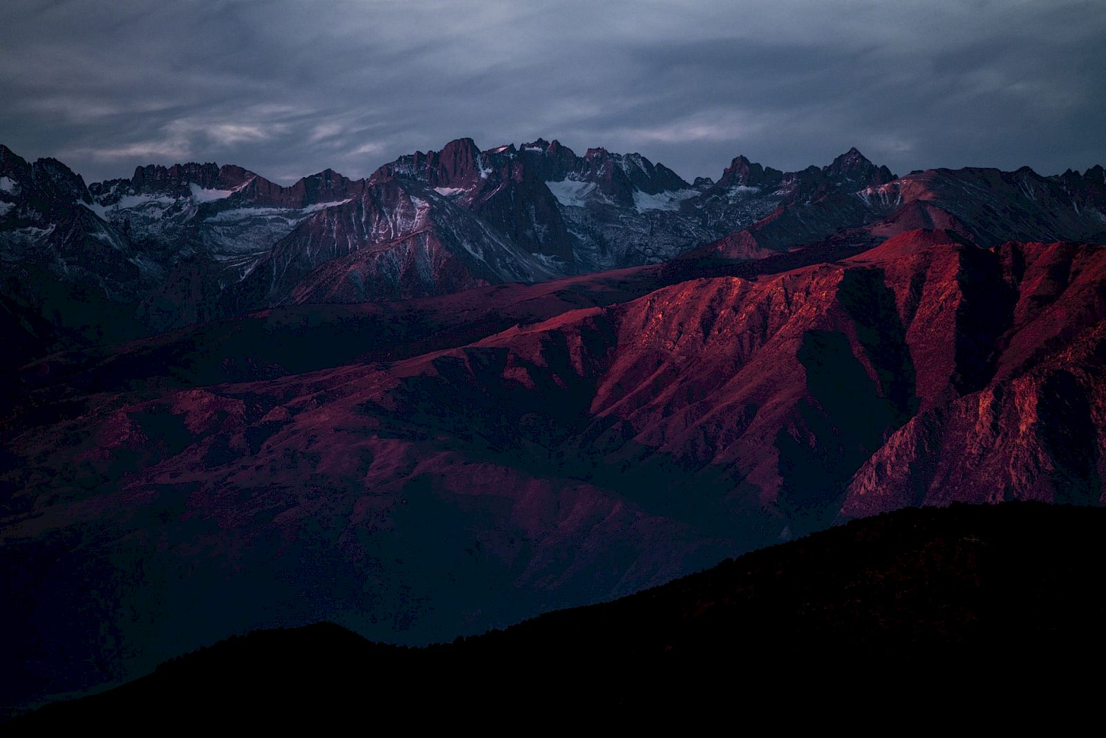 A mountain range at sunset with peaks bathed in reddish light and a dramatic sky in the background.