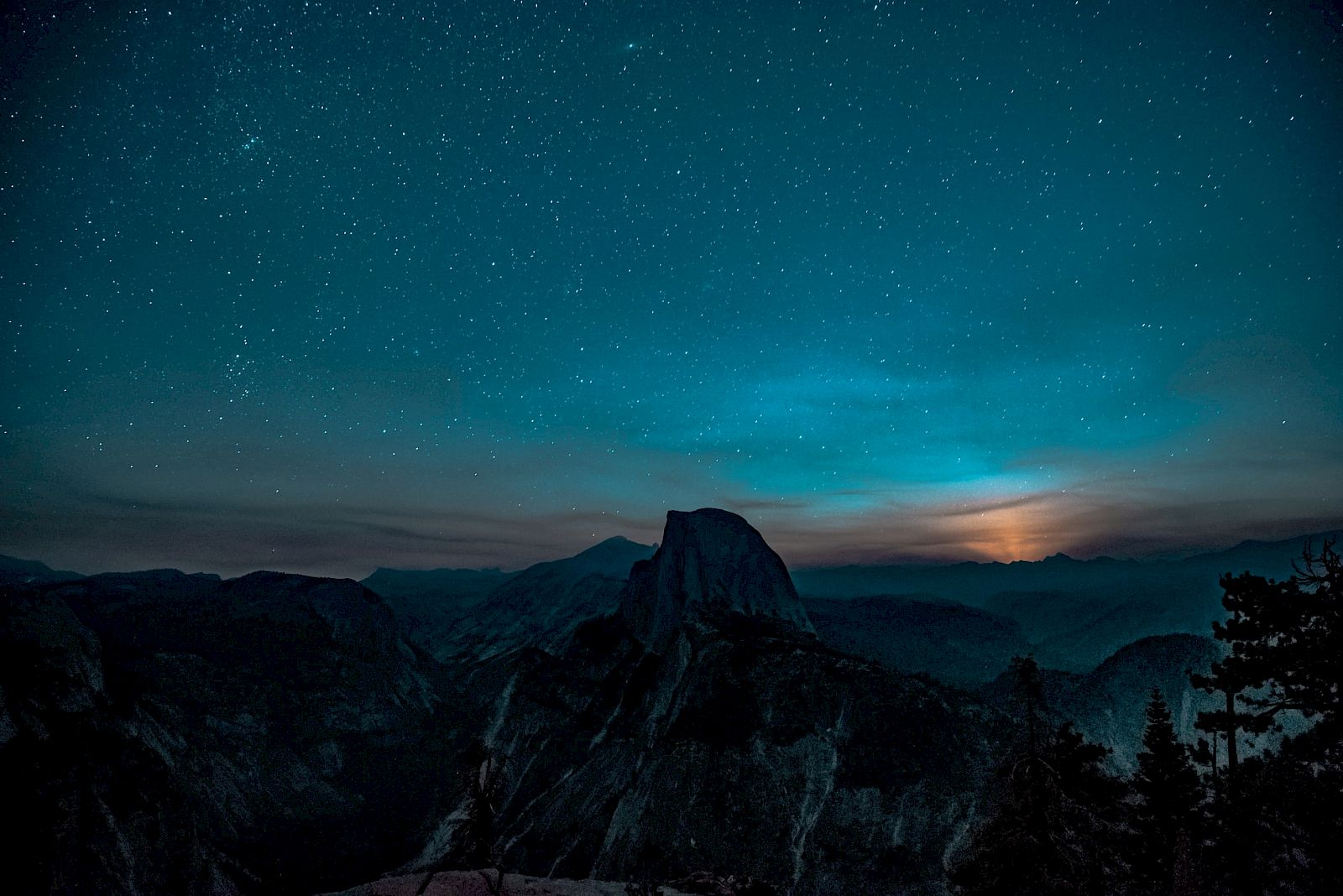 A starry night sky over a mountainous landscape with a silhouette of a prominent peak and a glowing horizon.