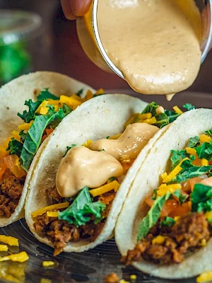 Three tacos with ground meat, lettuce, tomatoes, cheese, and a creamy sauce being poured on top on a glass plate.