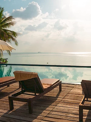 A tranquil poolside scene overlooking a calm sea, with sun loungers and umbrellas under a partly cloudy sky, surrounded by palm trees.