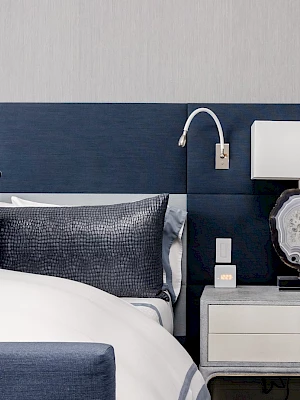 A modern bedroom with a blue bed, decorative pillows, and a nightstand featuring a lamp and a wall-mounted reading light.
