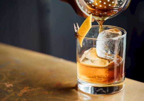 A hand pours a cocktail into a glass with a large ice cube and a citrus peel garnish on a wooden surface.