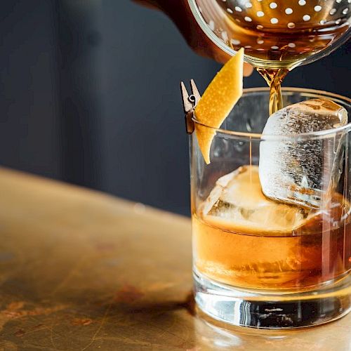 A hand pours a cocktail into a glass with a large ice cube and a citrus peel garnish on a wooden surface.