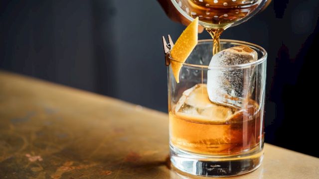 A hand pours a cocktail into a glass with a large ice cube and a citrus peel garnish on a wooden surface.