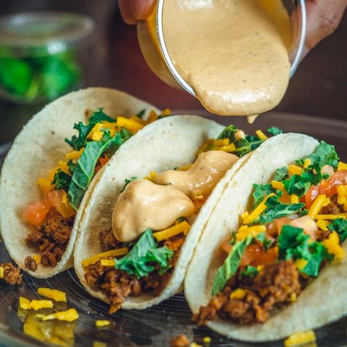 Three tacos filled with meat, shredded cheese, lettuce, and tomato, topped with a creamy sauce being poured over them on a plate.
