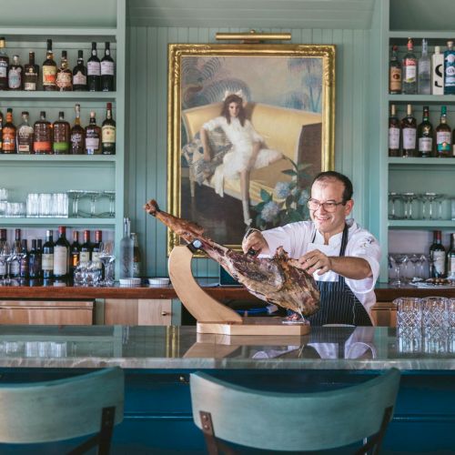 A chef slices cured meat at a bar with a large selection of bottles and a painting of a dog in the background.