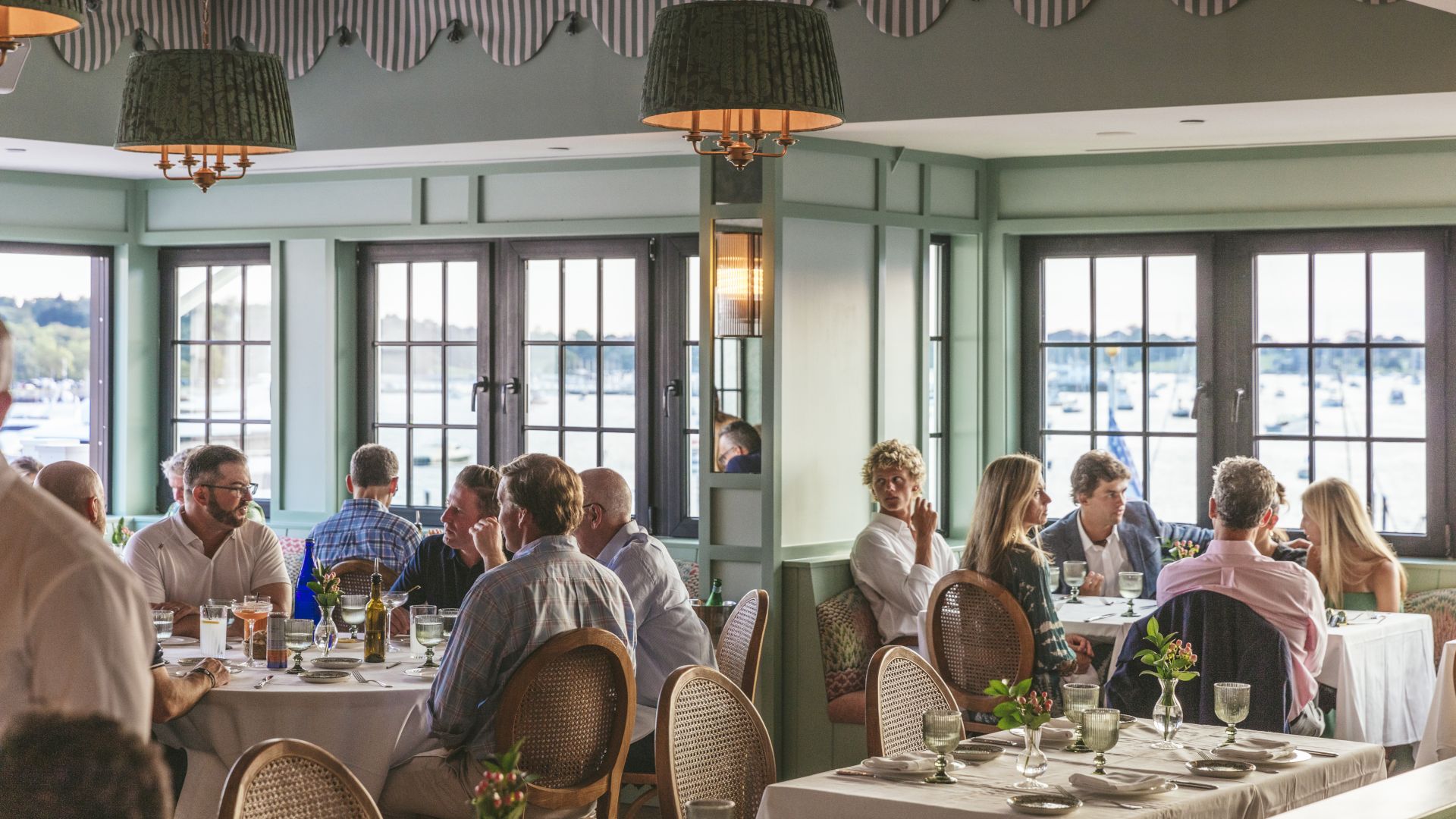 People dining in a restaurant with large windows showing an outdoor view. Tables are set with glassware and plates.