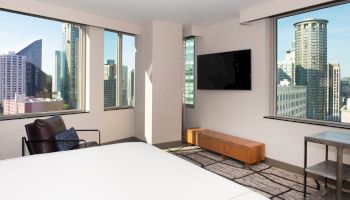 A modern hotel room with large windows shows a cityscape view, featuring a bed, wall-mounted TV, chair, and small table with a lamp.