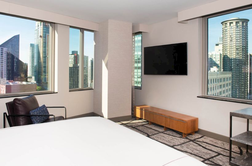 A modern hotel room with large windows shows a cityscape view, featuring a bed, wall-mounted TV, chair, and small table with a lamp.