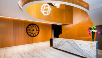 A modern lobby with a marble reception desk, wooden walls, a large clock, decorative lighting, and a vase of purple and white flowers.