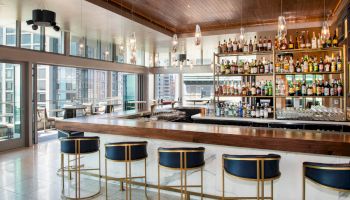 A stylish bar with high-back chairs, shelves stocked with bottles, and large windows offering a city view in a modern, well-lit setting.