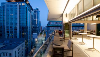 A modern rooftop patio with tables and chairs overlooking a cityscape with tall buildings at dusk.