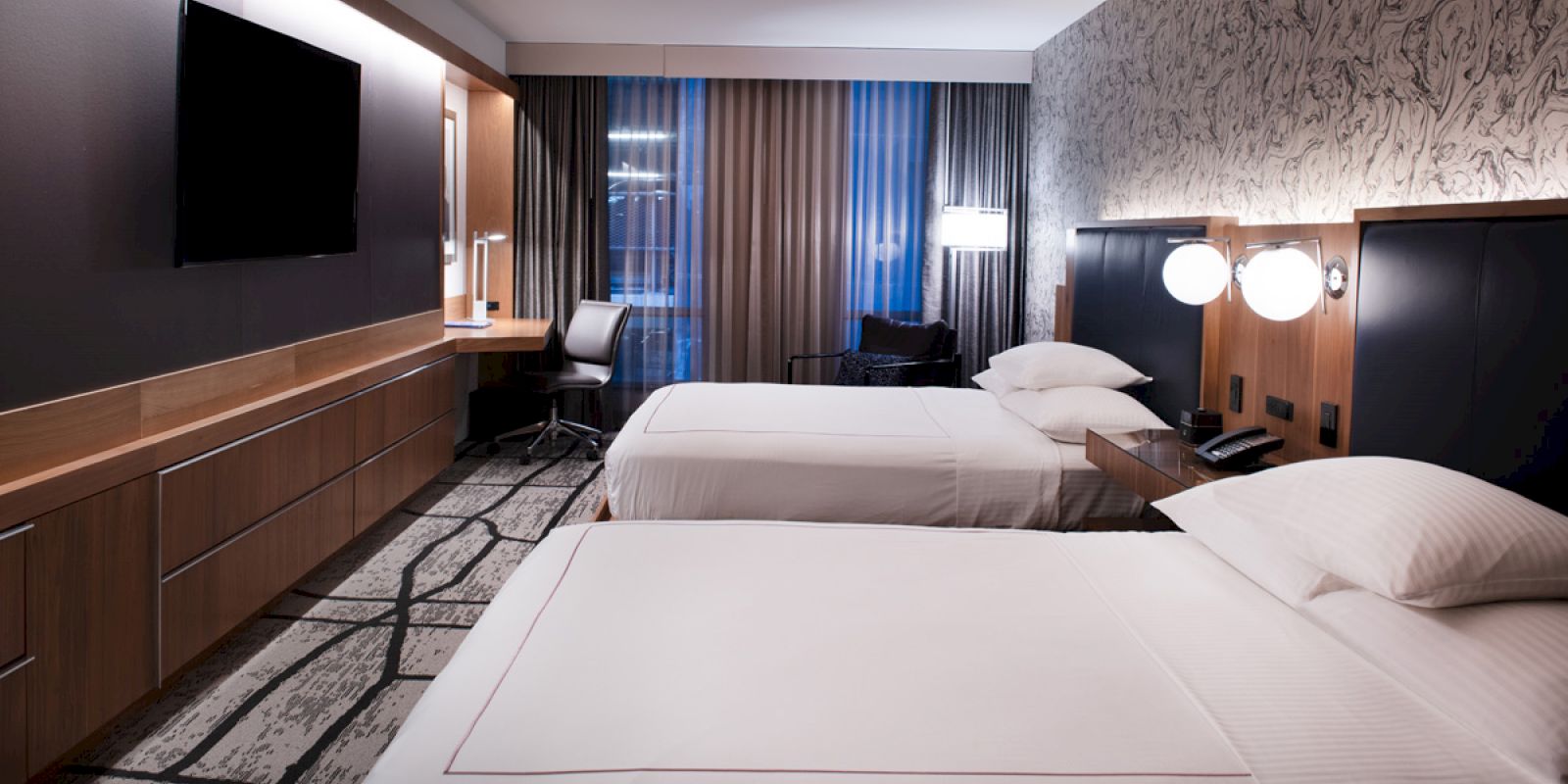 A modern hotel room with two beds, a desk and chair, a large TV, patterned carpeting, and stylish lighting on a decorated feature wall.