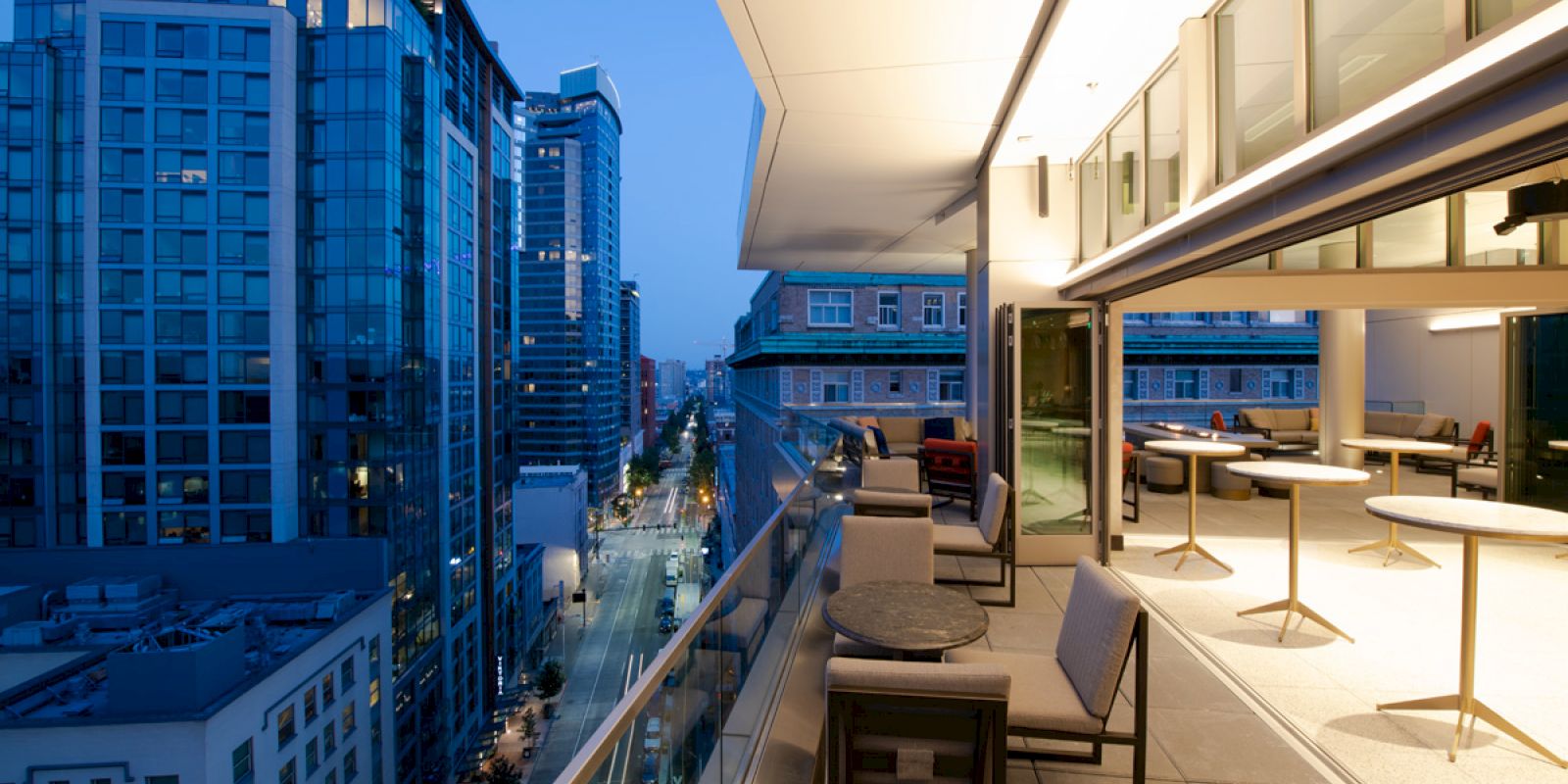 A modern rooftop terrace with seating overlooks a city street, surrounded by tall buildings at dusk, illuminated by soft lighting.