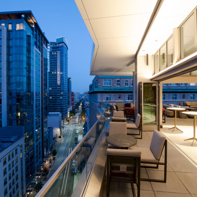 A modern rooftop terrace overlooking a cityscape with tall buildings and street view at dusk, featuring seating and tables.