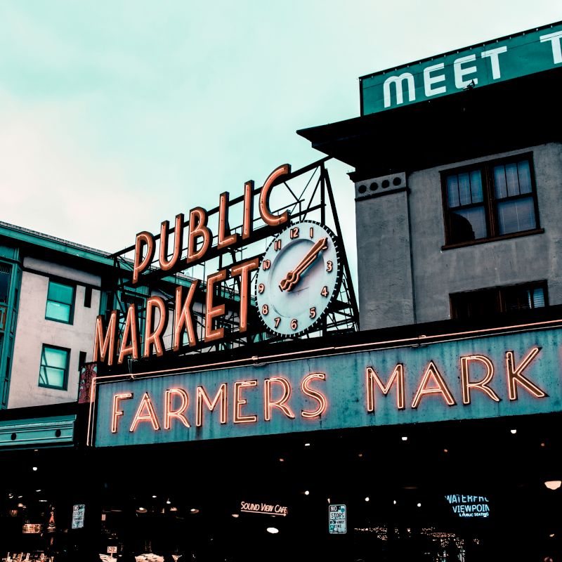 The image shows a neon sign for a "Public Market" and "Farmers Market" with a clock, set against the backdrop of urban buildings.