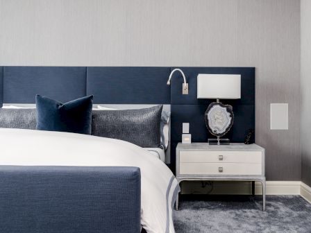A modern bedroom with a blue headboard, neatly made bed, white bedside table, lamp, decorative stone piece, and wall-mounted reading light.
