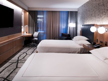The image shows a modern hotel room with two beds, a TV, desk, chair, and patterned carpet, featuring stylish lighting and decor.