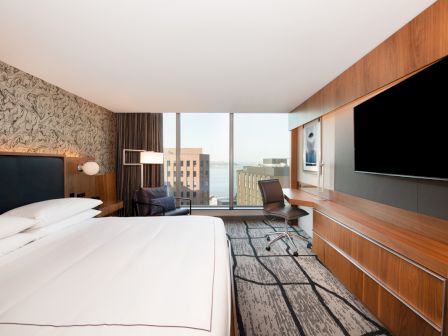 A modern hotel room with a large bed, desk, chair, and a flat-screen TV, featuring a cityscape view through a large window.